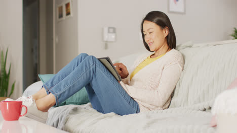 Video-of-happy-asian-woman-resting-on-sofa-with-tablet