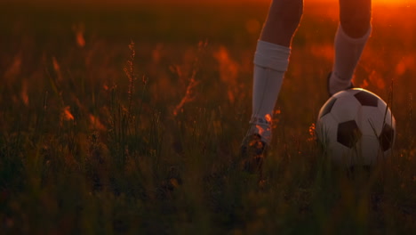 close-up of tracking the feet of a football boy in a red t-shirt and shorts running with the ball at sunset in the field on the grass. the young football player dreams of a professional career and trains in the field. on the way to success