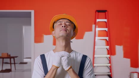 un pintor asiático que usa un casco de seguridad orando por algo mientras está de pie con la pared de la pintura en la habitación