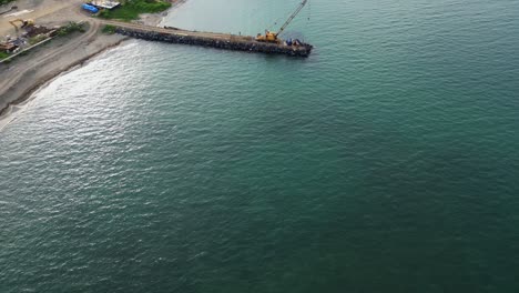 aerial look-up drone shot of coastal breakwater construction site and yellow crane at tropical island during daytime.