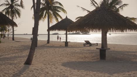 Mother-with-her-child-walking-together-on-a-beach-behind-palm-trees-during-summer-sunset-warm-golden-hour-on-tropical-island