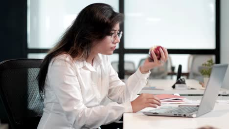 a beautiful and health conscious girl wearing eyeglasses taking a bite of an apple while working on a laptop on a creative project.