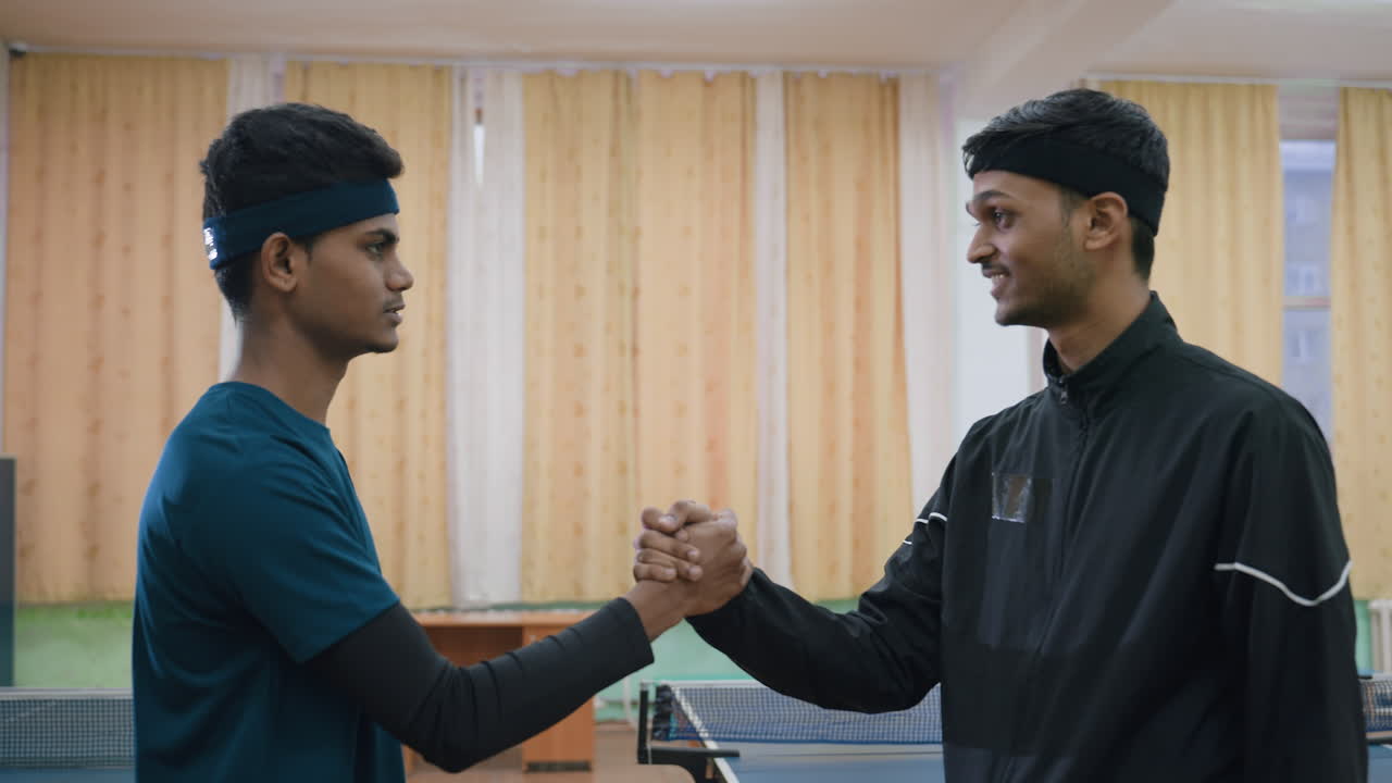 Two athletes shake hands near tennis table indoors, one engaging with friendly expression while other looks serious, capturing sportsmanship, respect, and interaction in competitive training
