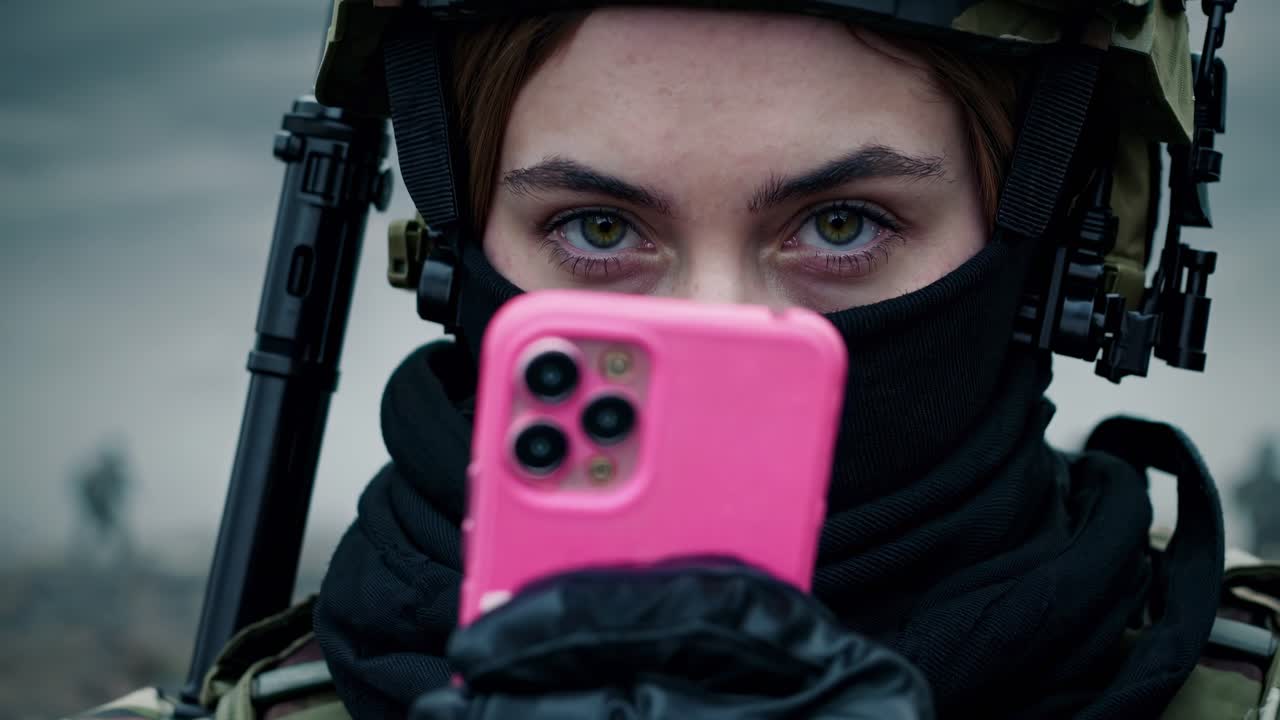 Female soldier wearing balaclava and holding pink smartphone, with assault rifle visible in background, during military operation in war zone