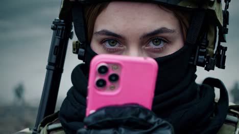 female soldier wearing balaclava and holding pink smartphone, with assault rifle visible in background, during military operation in war zone
