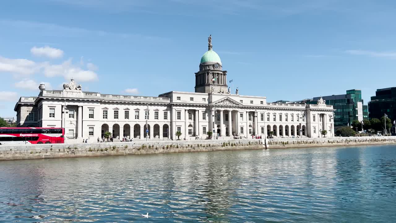 View across the Liffey River onto the southern facade of Custom House in Dublin City on a sunny day with blue skies. There are a lot of cars, buses, and people on the streets. Handheld shot