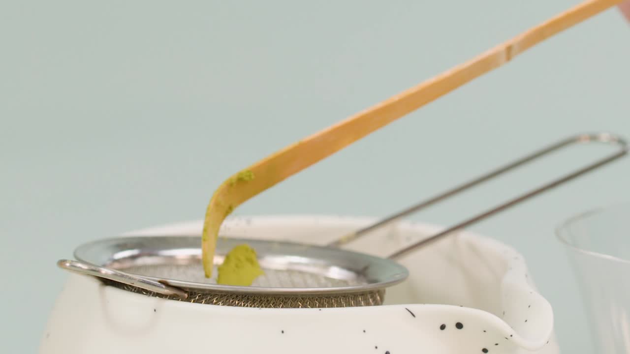 Green tea powder falls from a bamboo spoon into a metal strainer over a bowl