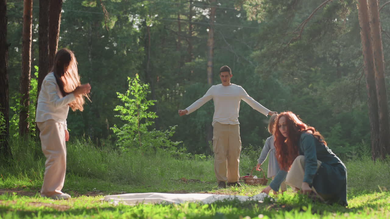 Teenage girl and mom spreading blanket on grass while siblings watch nearby, warm sunlight shining through forest trees creating peaceful family picnic moment full of cooperation, joy, and love