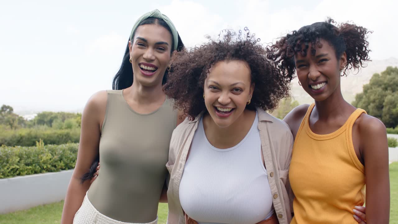 Amigas diversas posando para a câmera, usando blusas, sorrindo e rindo, compartilhando alegria no parque.