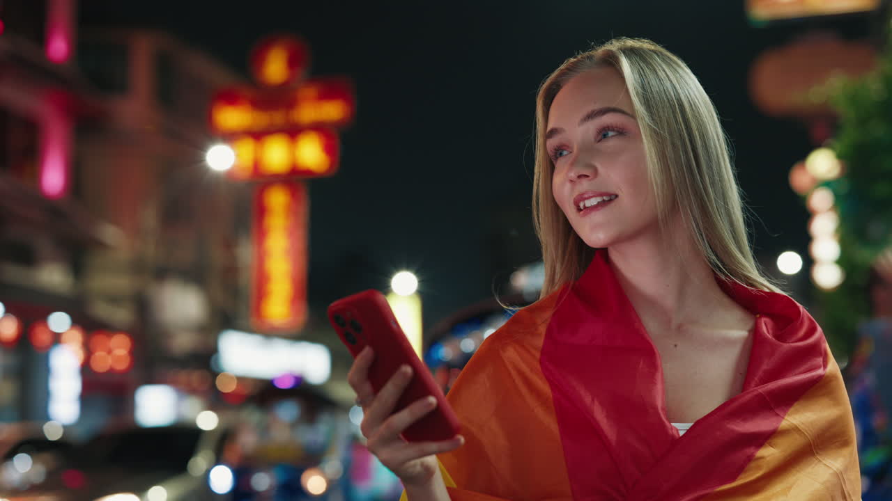 Donna che usa il telefono in una strada cittadina di notte, con una sciarpa con la bandiera Pride
