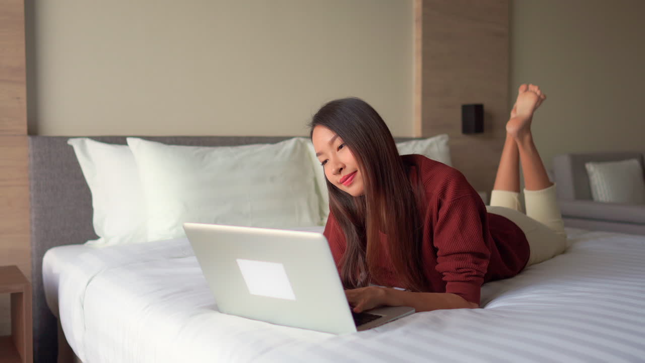 젊고 매력적인 아시아 여성이 노트북과 상호 작용할 때 나쁜 자세로 누워 있는 동안 미소를 짓습니다.
