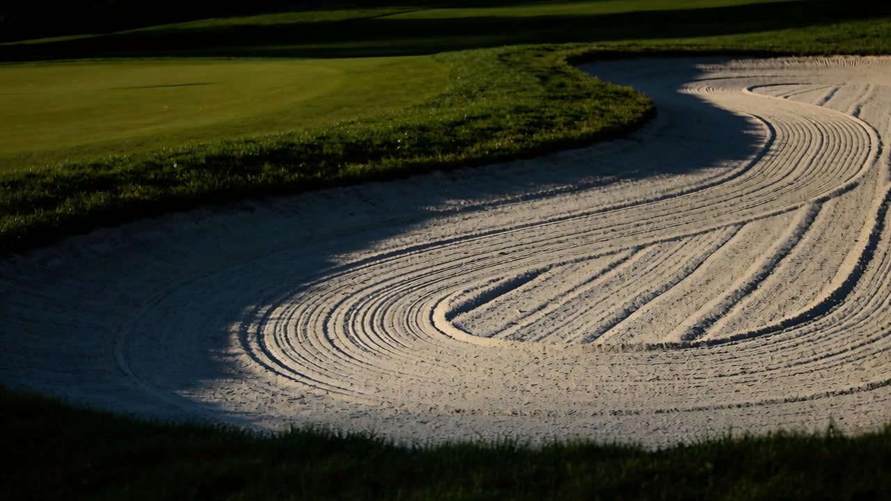 Kamera zoomt und enthüllt einen geharkten Sandbunker auf einem Golfplatz, um Rillen, Putting Green und Schatten zu zeigen.