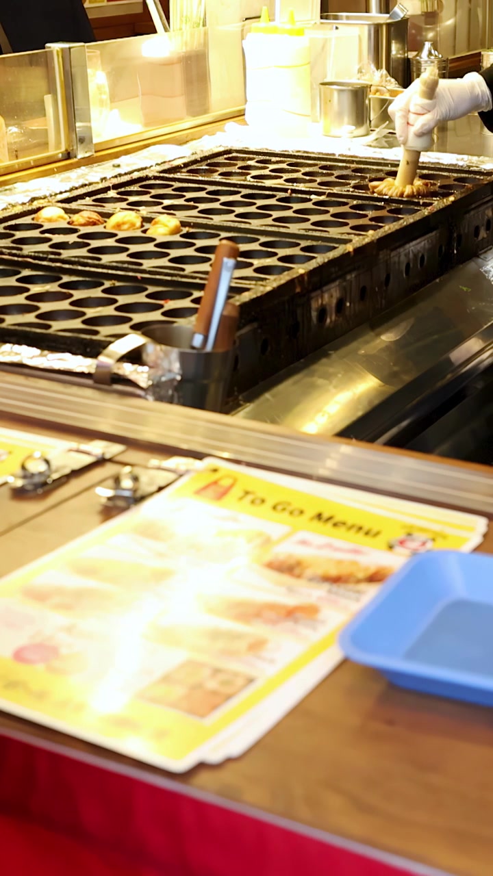 En kokk med hansker bruker et treredskap til å lage blekksprutboller på en varm grill
