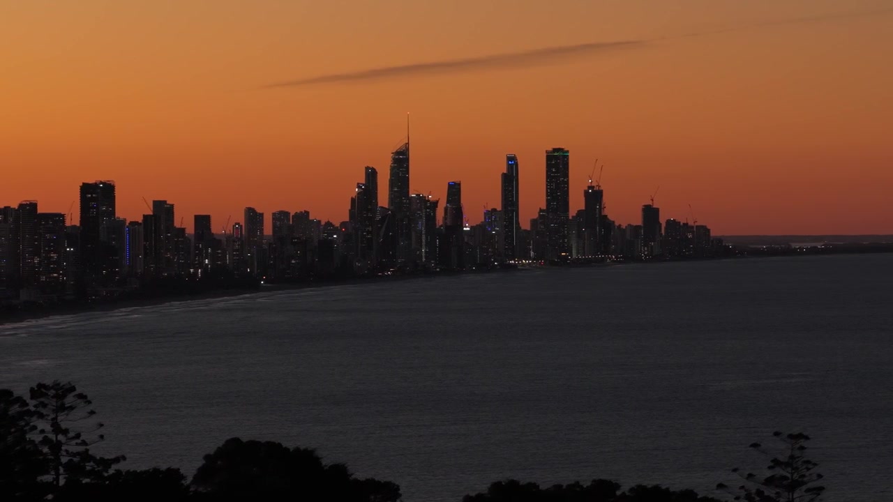 Toma aérea lateral con dron moviéndose de izquierda a derecha a través del horizonte de Surfers Paradise y Broadbeach al anochecer desde Burleigh Headland en Gold Coast, Queensland, Australia.