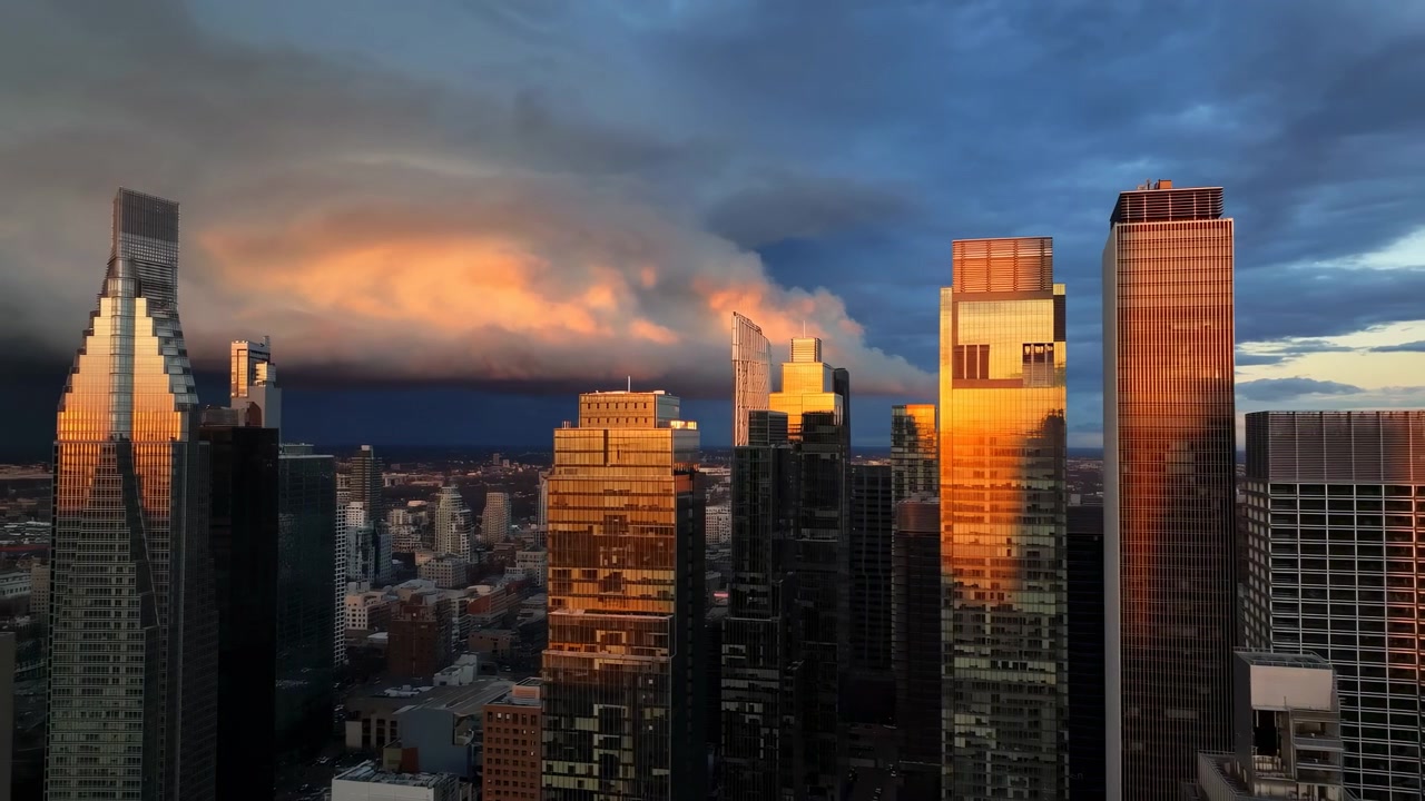 Vista lateral lenta revela fachadas iluminadas de arranha-céus e ruas sombrias, enquanto bordas de nuvens alaranjadas se estendem sobre a cidade, contrastando reflexos dourados com um céu tempestuoso e frio
