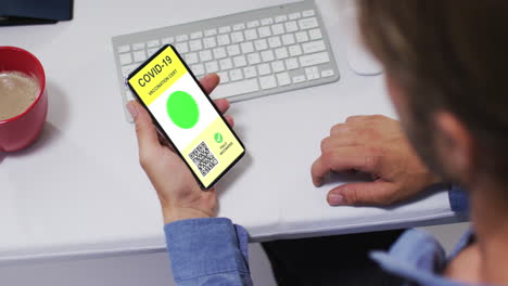 hombre en el escritorio sosteniendo un teléfono inteligente con certificado de vacunación contra la covid y código qr en la pantalla
