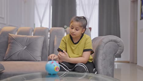 Sad-Indian-kid-girl-putting-coins-in-piggy-bank