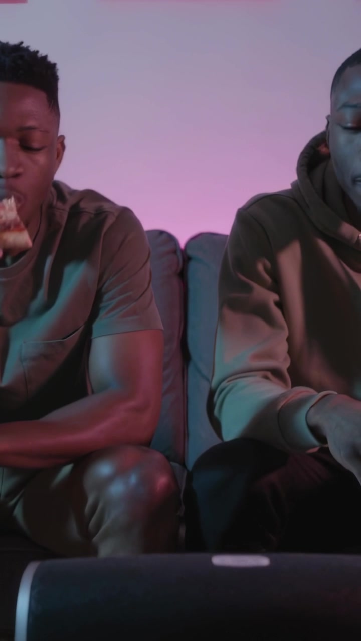 Vertical video: Panning men on sofa, left tee snacking on pizza, right hoodie using phone, speaker
