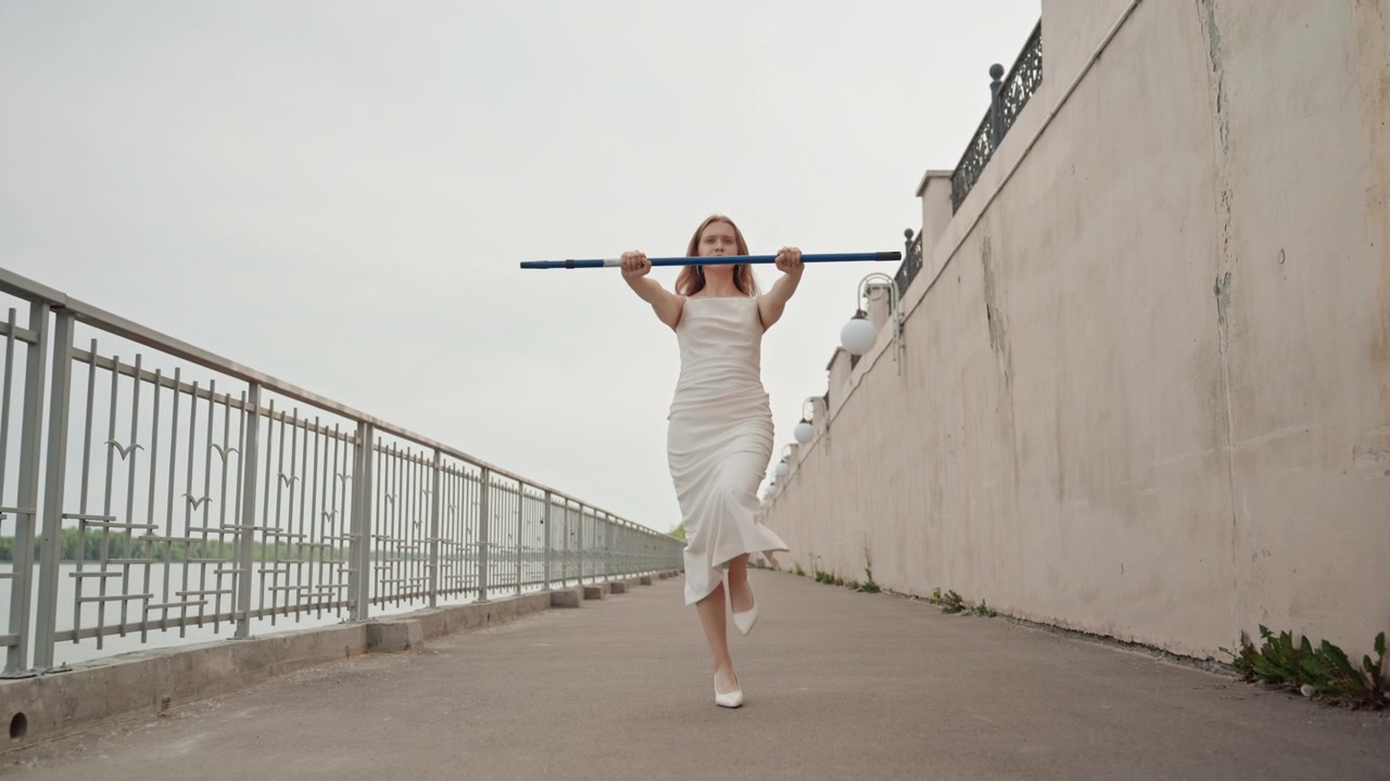 Una donna in abito bianco cammina lungo un sentiero all'aperto tenendo un palo blu sopra la testa. La scena presenta ringhiere metalliche, muri di cemento e acqua distante sotto una luce nuvolosa con ombre tenui.
