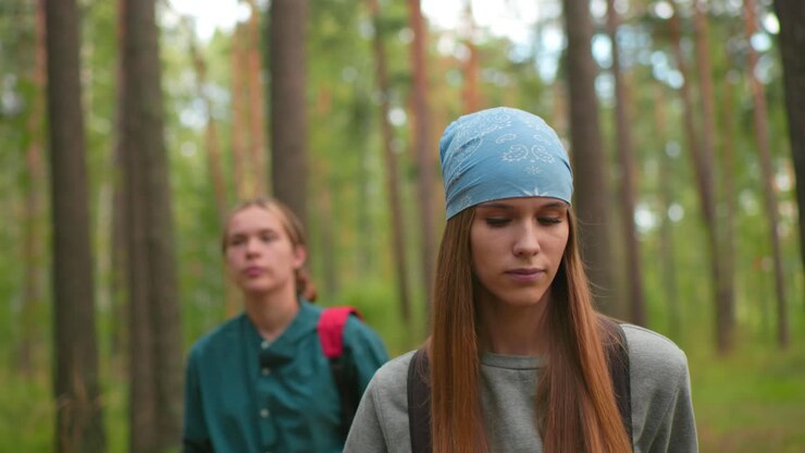 Women hiking along forest trail, woman in gray sweatshirt appears tired, gazing forward determinedly, while her companion in green shirt glances around, softly blurred in background