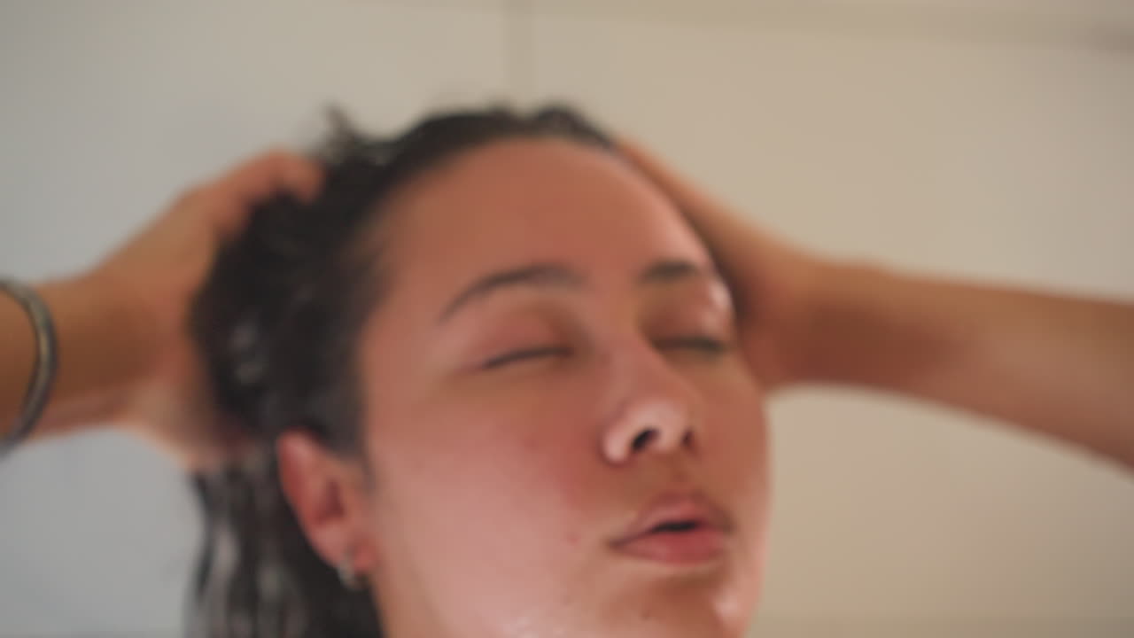 Donna ospite nella doccia di un hotel, occhi chiusi mentre si sciacqua i capelli, goccioline d'acqua che brillano sulla pelle. Scena intima di un bagno privato con asciugamano e rubinetto visibili, atmosfera calma da spa e vapore delicato, rilassata.