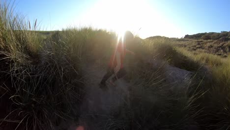 Vista-Trasera-Del-Joven-En-Vacaciones-En-La-Playa-Corriendo-A-Través-De-Dunas-De-Arena-Contra-El-Sol-Abrasador