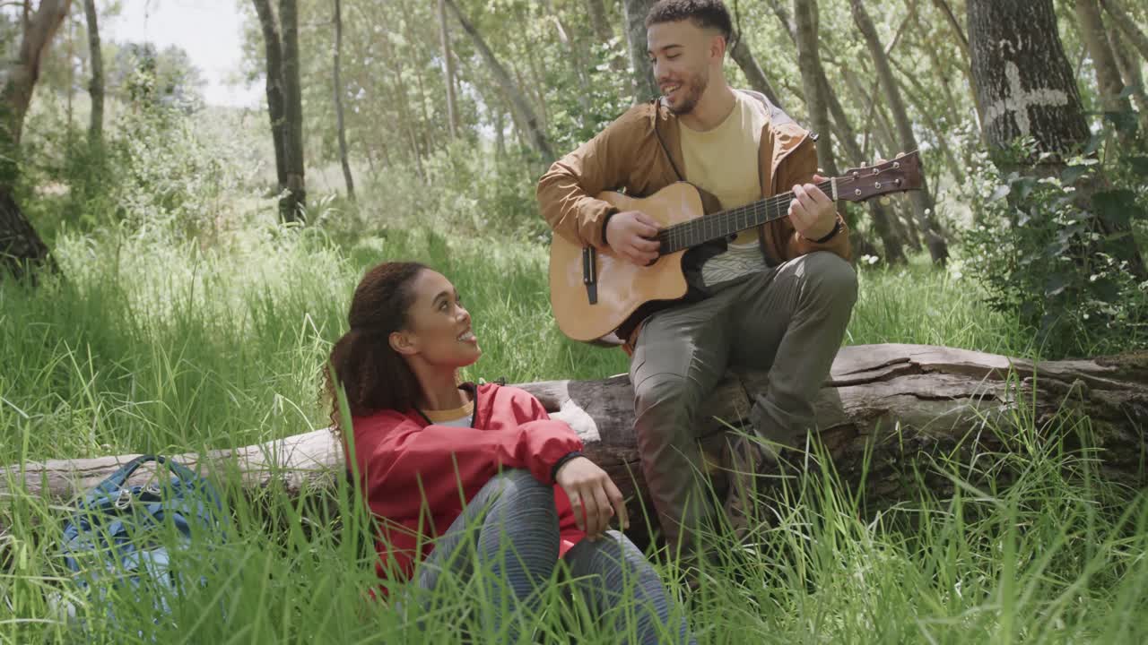 feliz pareja afroamericana sentada en el tronco de un árbol y tocando la guitarra en el bosque, cámara lenta
