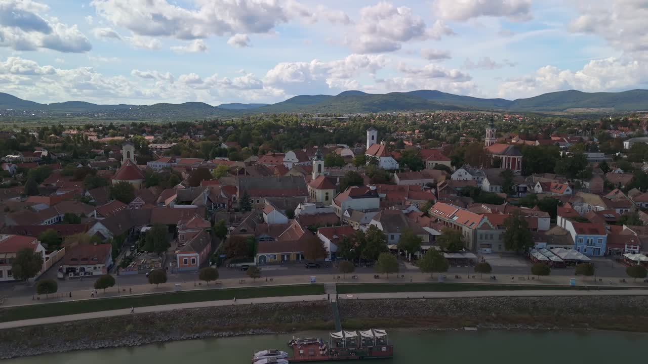 Drone-opptak sidelengs som beveger seg mot høyre over Szentendre, Ungarn, og fanger de fargerike hustakene, barokke kirkene og strandpromenaden ved elven mot det naturskjønne bakteppet av Pilis-fjellene