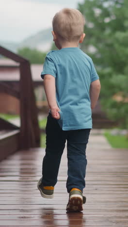 playful blond toddler boy runs holding t-shirt edge along veranda deck at modern cottage house after warm spring rain side view slow motion
