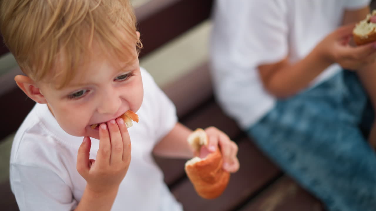 A child is intently eating a sausage roll, using his hand to push the food into his mouth, another child is visible in the background, slightly blurred