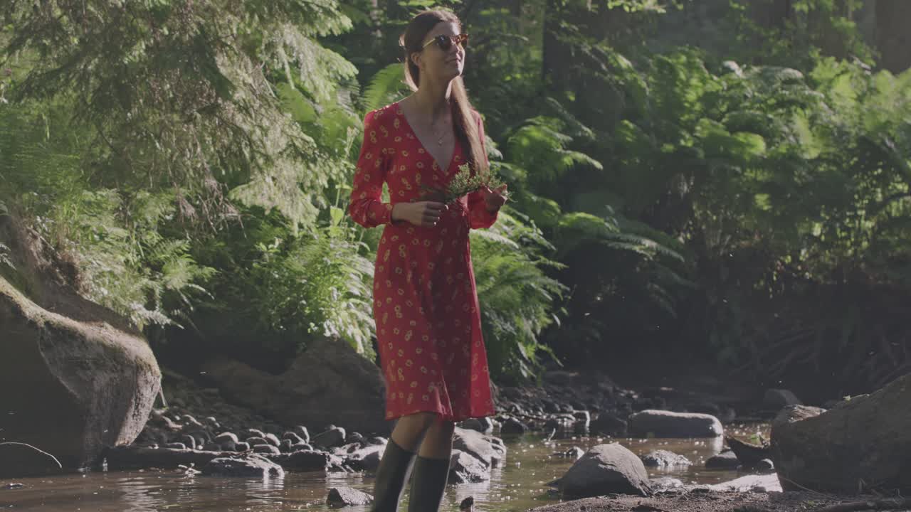 Enchanted Forest Stroll: Camera pans up from a water reflection of a woman in a vibrant red dress holding a juniper branch as she walks through mist and fog in a fairy-tale forest