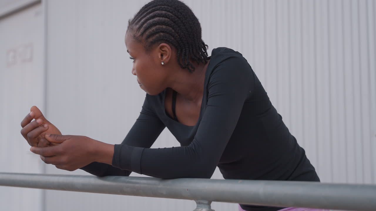 black teen leaning on railing, pensive gaze and braided hair against industrial wall, evening light, athlete taking a break after training, introspective mood and soft depth of field