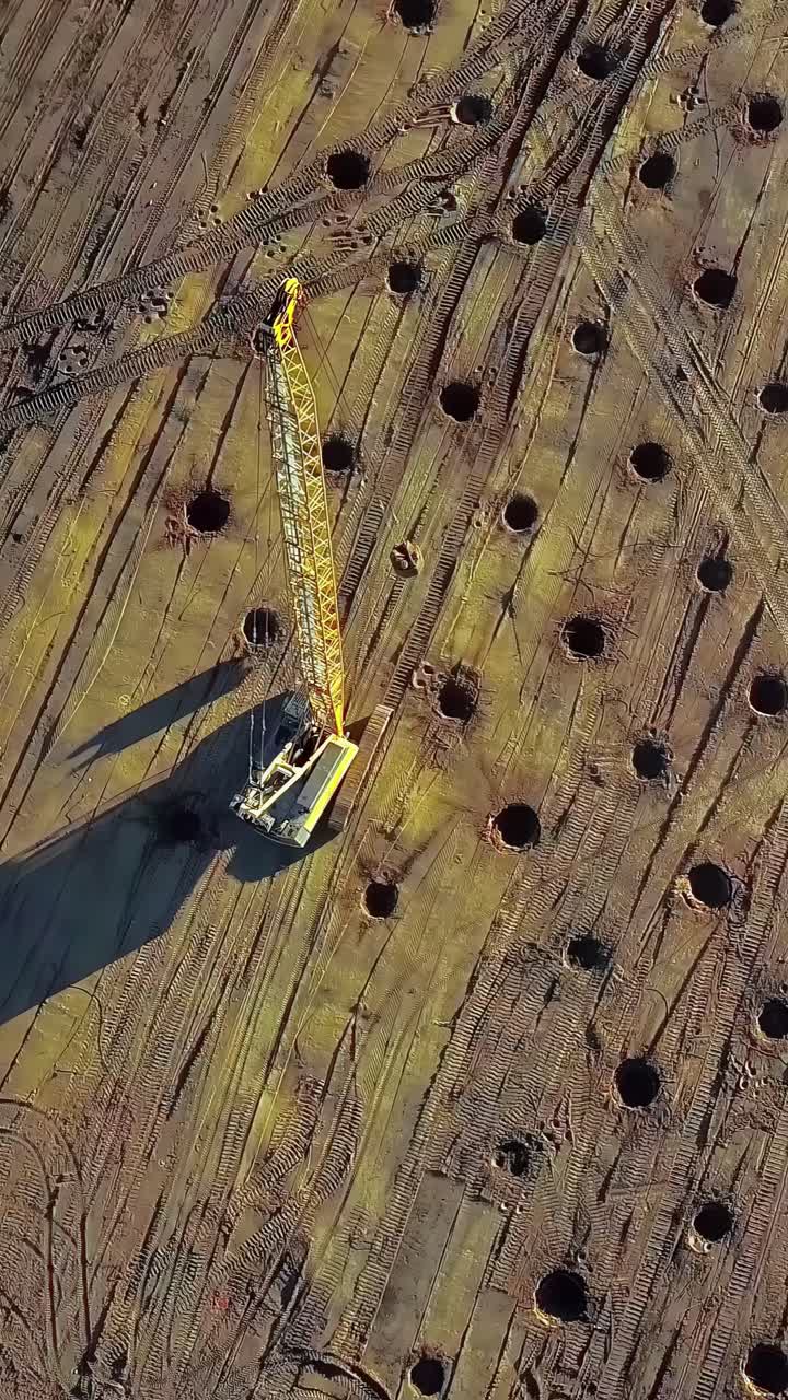 A top-down aerial parallax shot shows a large crawler crane at a construction site in Latvia, with a grid of deep holes dug for the pile foundations of a new building or structure