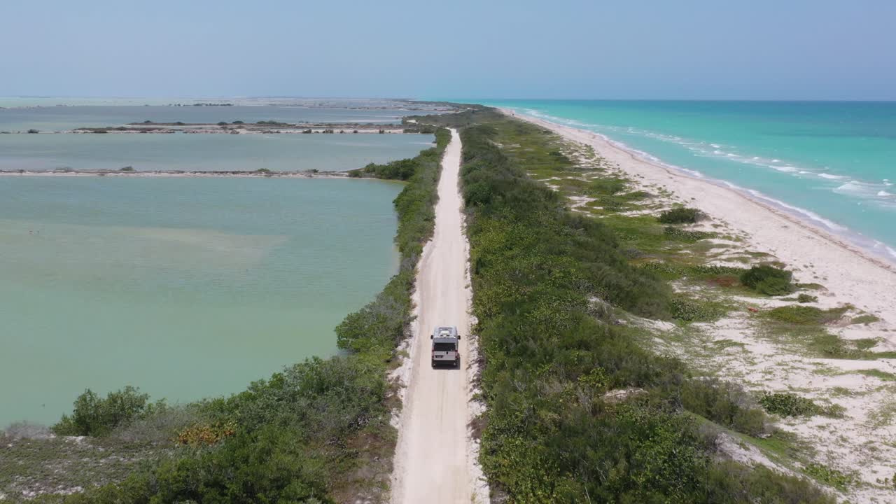 Following drone shot of Camper Van driving white sandy road between turquoise Caribbean ocean and green salt lake view. Mexico, Rio Lagartos