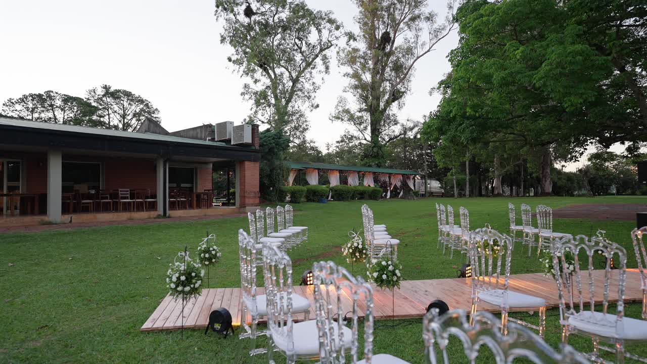Allestimento per cerimonia nuziale all'aperto su un prato, con una casa per eventi e alberi alti sullo sfondo. Ripresa in slow motion che scorre da destra, nel pomeriggio.