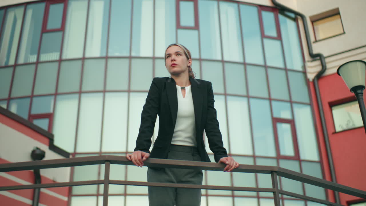 Professional girl in formal outfit leans on iron railing, gazing into distance with focused expression, beside lamp post and CCTV camera, modern glass office building in background