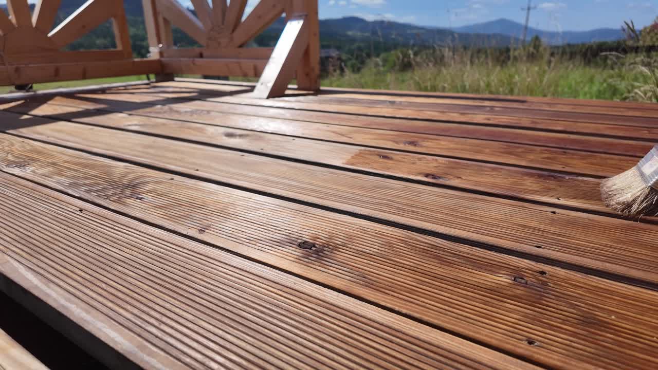 Hand oils and impregnate wooden terrace boards with brush. Decorative railing, serene sky, clouds, and mountain horizon evoke home improvement and outdoor maintenance themes