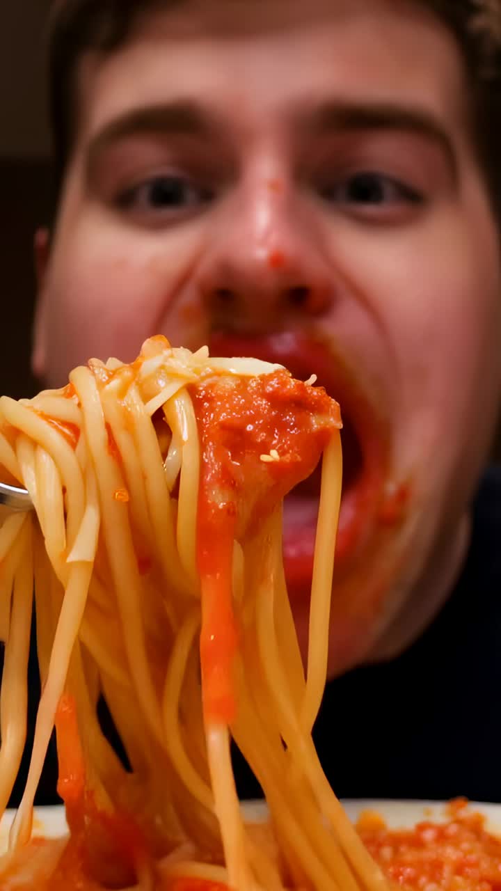 Video verticale: forchetta roteante, uomo spinto dalla fame che mangia spaghetti in cucina, sugo sulla camicia