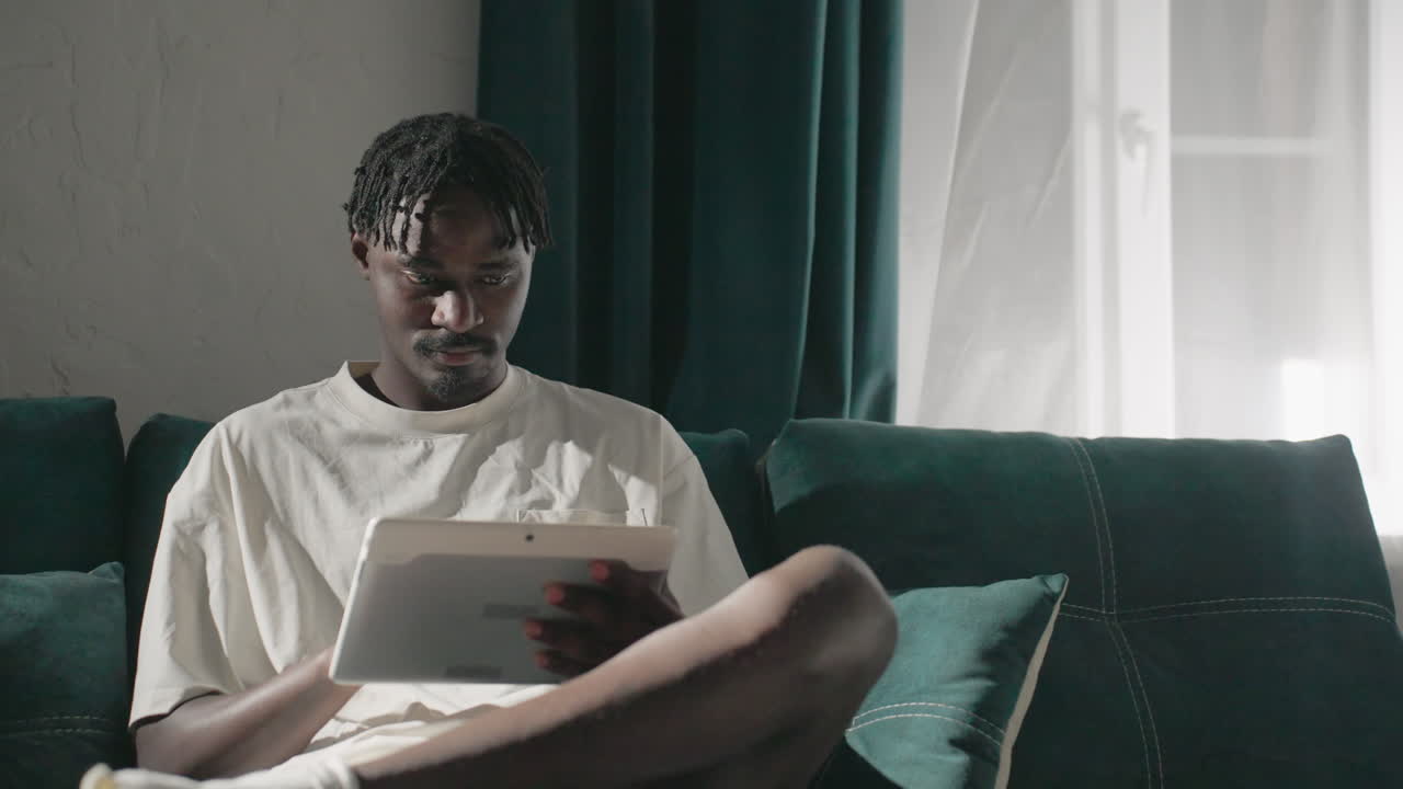 Champ focused on tablet, seated on teal sofa in cozy living room. Sunlight shines through window, creating a calm, peaceful atmosphere perfect for relaxation and personal time