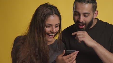 studio shot of excited couple with mobile phone celebrating winning money against yellow background