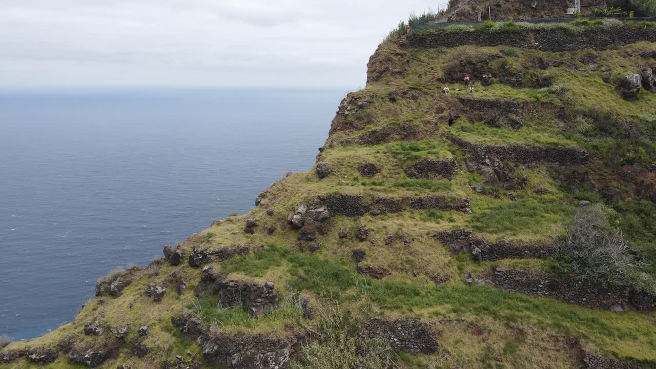 vista aérea de un acantilado empinado y en terrazas junto al océano con tres excursionistas en el terreno de hierba