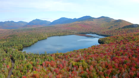 aerial view of a lake surrounded by beautiful fall colors with the high peaks of the adirondack mountains in the background, forward drone movement