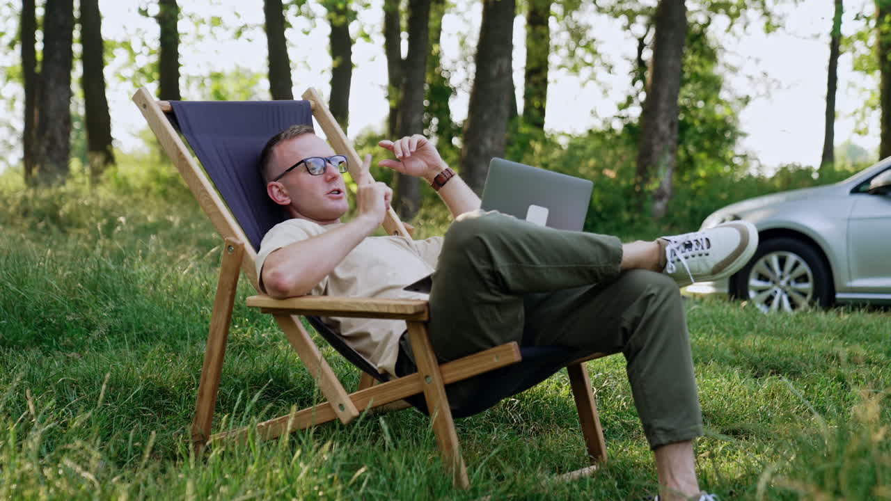 Relaxed Caucasian man sits in the folding chair. Businessman talk to someone online on laptop. Nature backdrop.