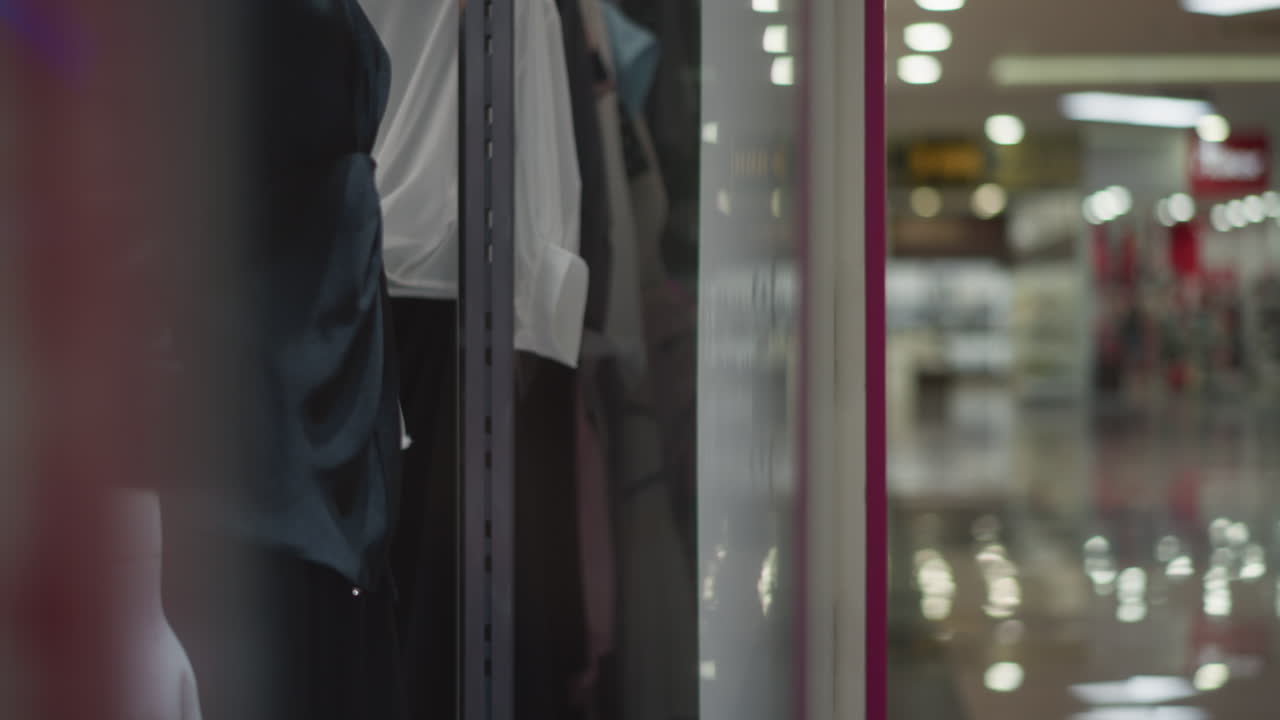 Close-up of clothing rack inside retail store with blurred reflection of bright mall interior, displays folded and hanging garments with soft focus and warm tones