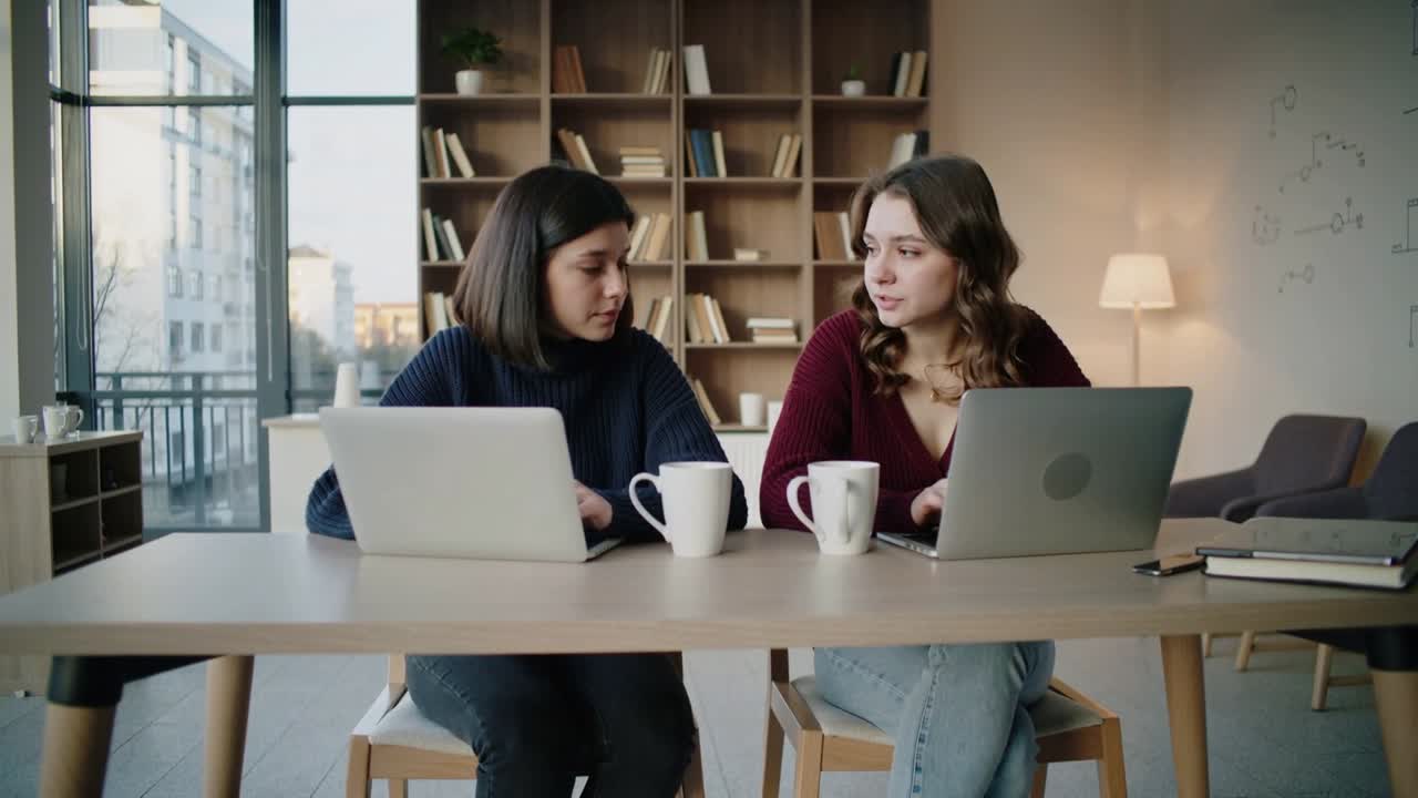 Two women working, right speaking and pointing at laptop, left adjusting and sipping for business