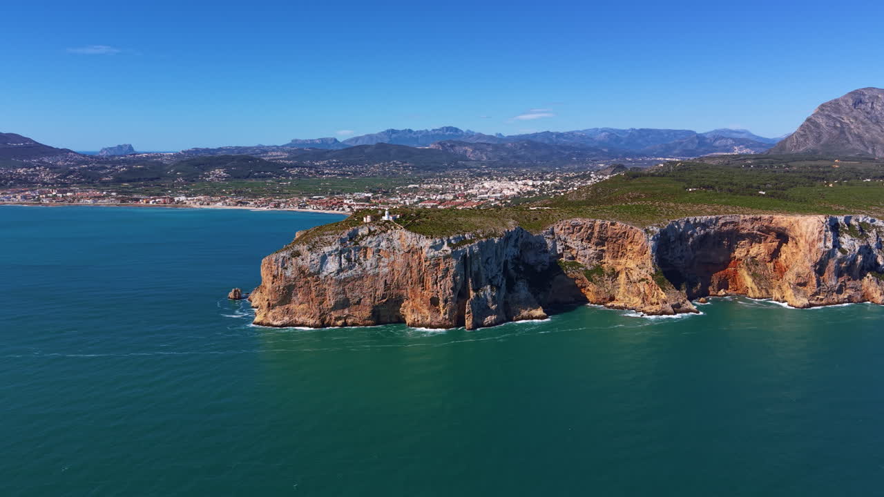 La penisola di Moraira sulla Costa Blanca, Spagna, con spettacolari scogliere calcaree che si ergono dalle acque turchesi del Mediterraneo, una città costiera e montagne sullo sfondo.