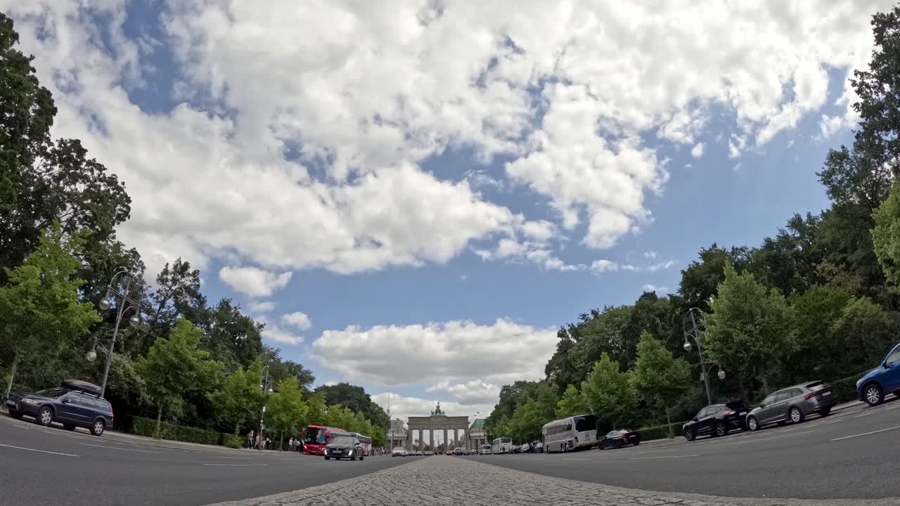 Low-angle timelapse captures cars driving past the Brandenburg Gate under partly cloudy skies, with lush green trees and dynamic daylight in Berlin