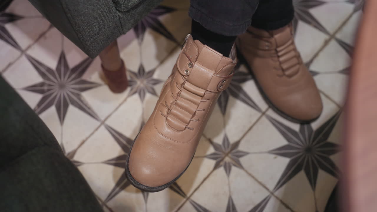 Close-up view of woman wearing brown leather boots with legs crossed and gently swinging over black and white patterned tile floor, creating relaxed indoor moment with stylish footwear and decor