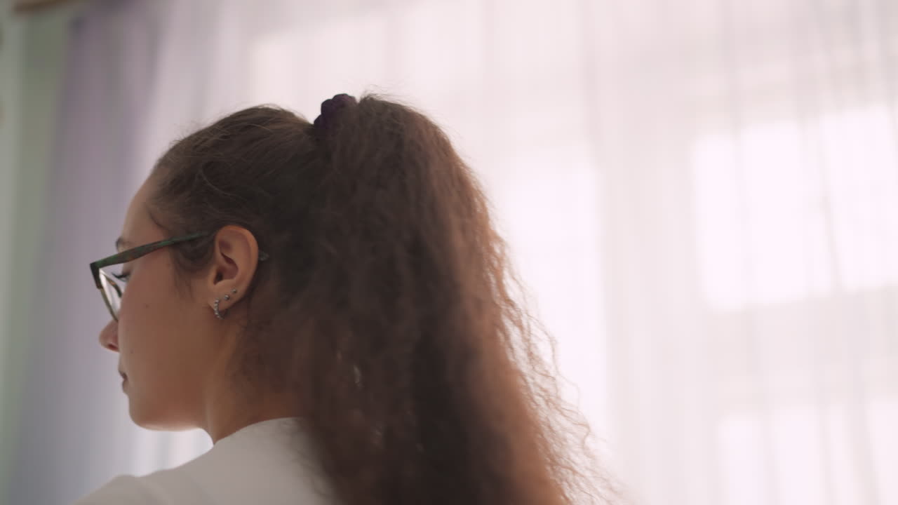 Close view of woman with curly brazil hair tied in ponytail wearing eyeglasses, side profile softly lit against curtain background, subtle indoor atmosphere showing detail of hairstyle, ear jewelry