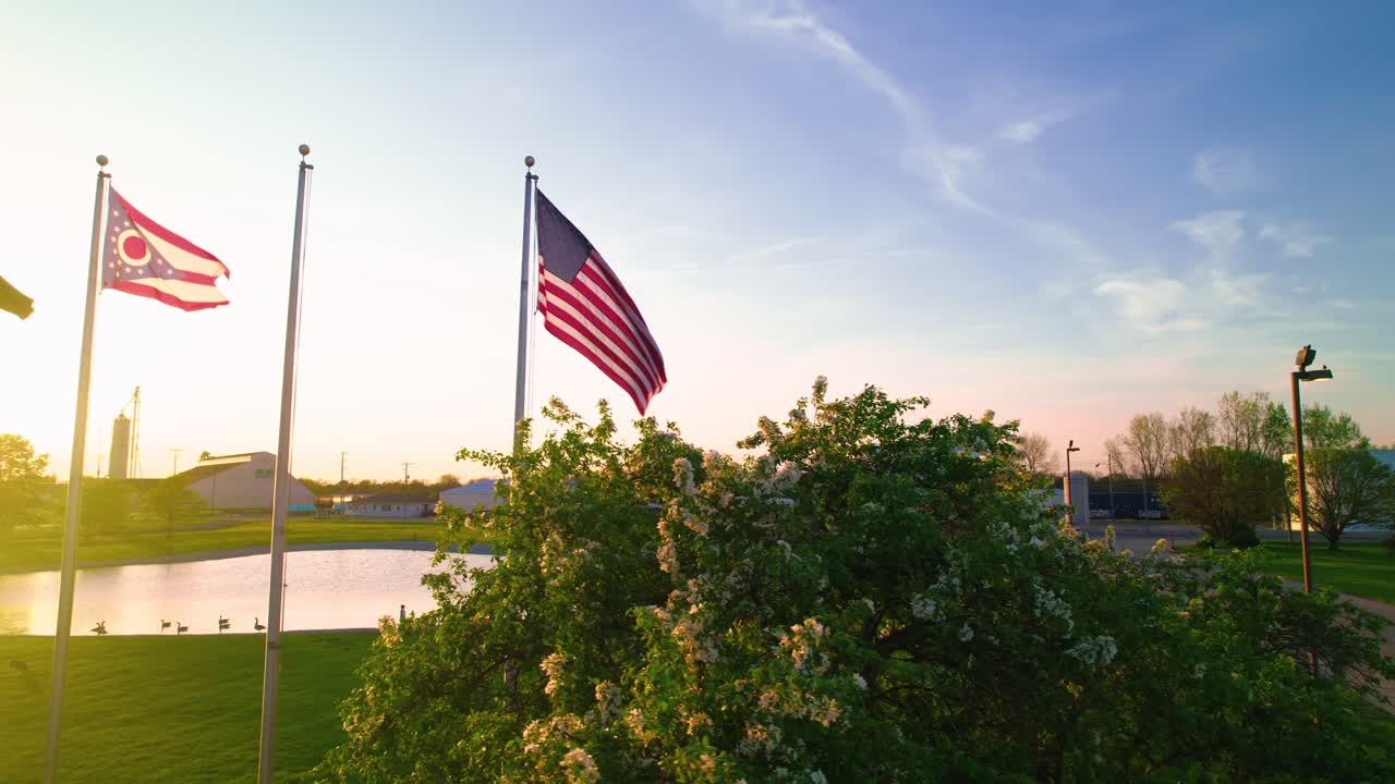 American and Ohio state flags waving at sunset beside industrial park and pond